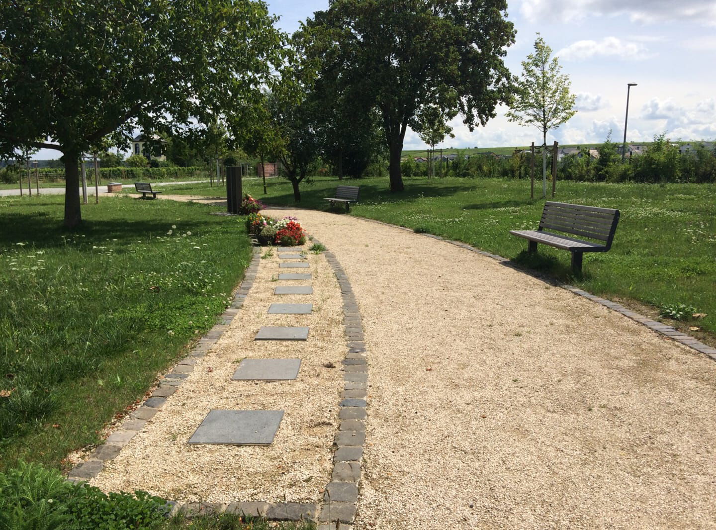 Friedhofsweg mit Sitzbänken, Bäumen und Blumen