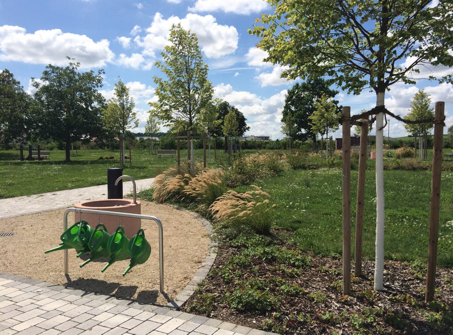 Brunnen mit Gieskannen und eine Wiese mit Bäumen im Hintergrund